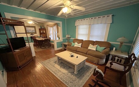 A living room with a sofa, coffee table, and a television on a stand. There is a dining area with a table and chairs, and a ceiling fan.