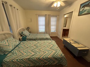 Une chambre avec deux lits, une commode, un ventilateur de plafond, une fenêtre avec des stores et un tableau au mur.