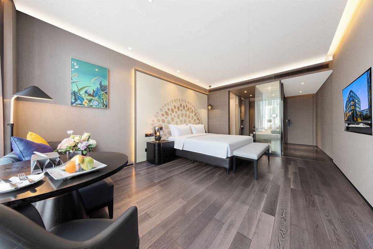 A modern hotel room with a bed, bedside table, lamp, and a dining area with a table and chairs.