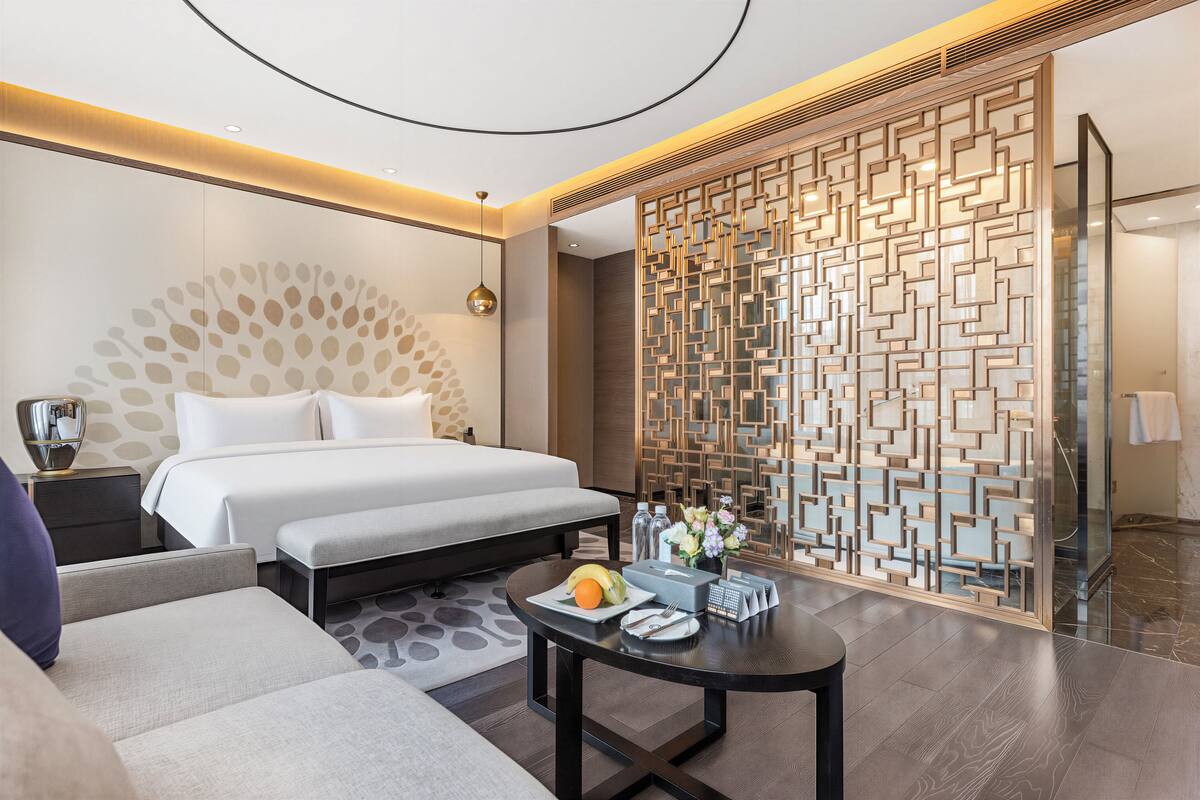 A hotel room with a bed, bedside table, a small round table with a fruit bowl, and a decorative partition.