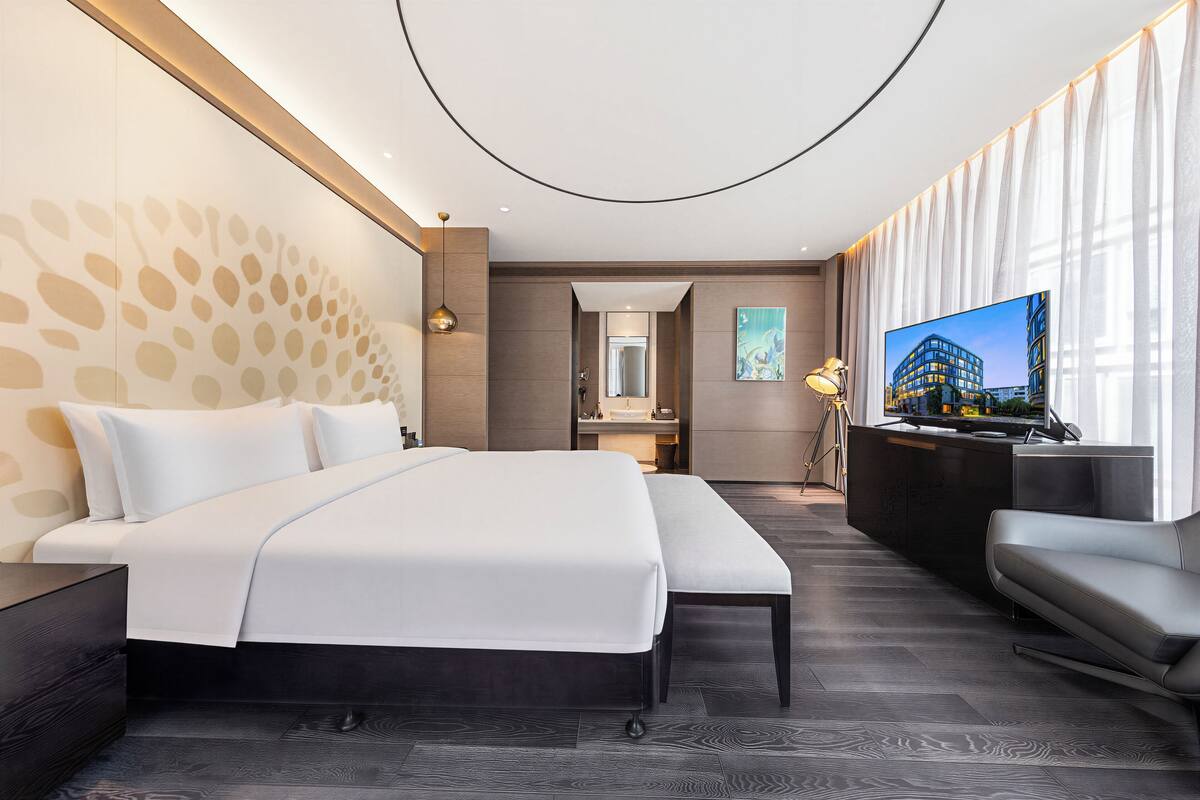 A modern hotel room with a large bed, a seating area, a TV, and a bathroom visible through an open door.