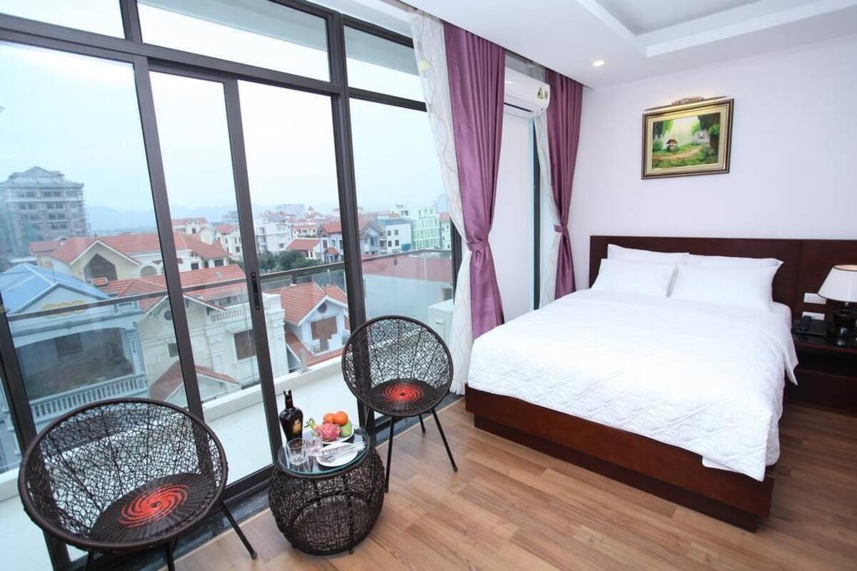 Habitación de hotel con una cama grande, dos sillas de mimbre, una mesita con una bebida y fruta, y vistas a la ciudad a través de grandes ventanales.