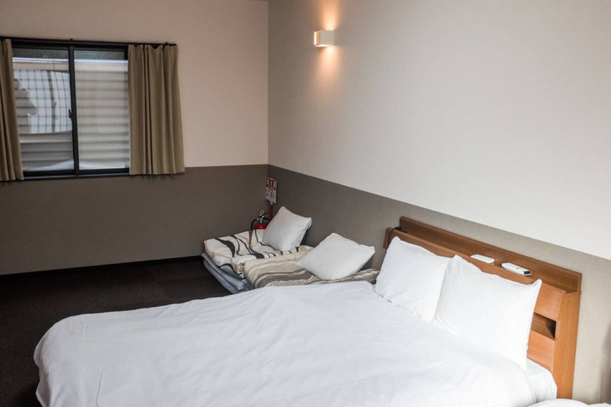 A hotel room with a large bed, a smaller bed, a window with curtains, and a wall-mounted light fixture.