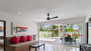 A modern living room with a sofa, coffee table, and a TV. There is a sliding glass door leading to a pool area with outdoor furniture. - SAWAN Pool Villas Residence (Koh Samui)