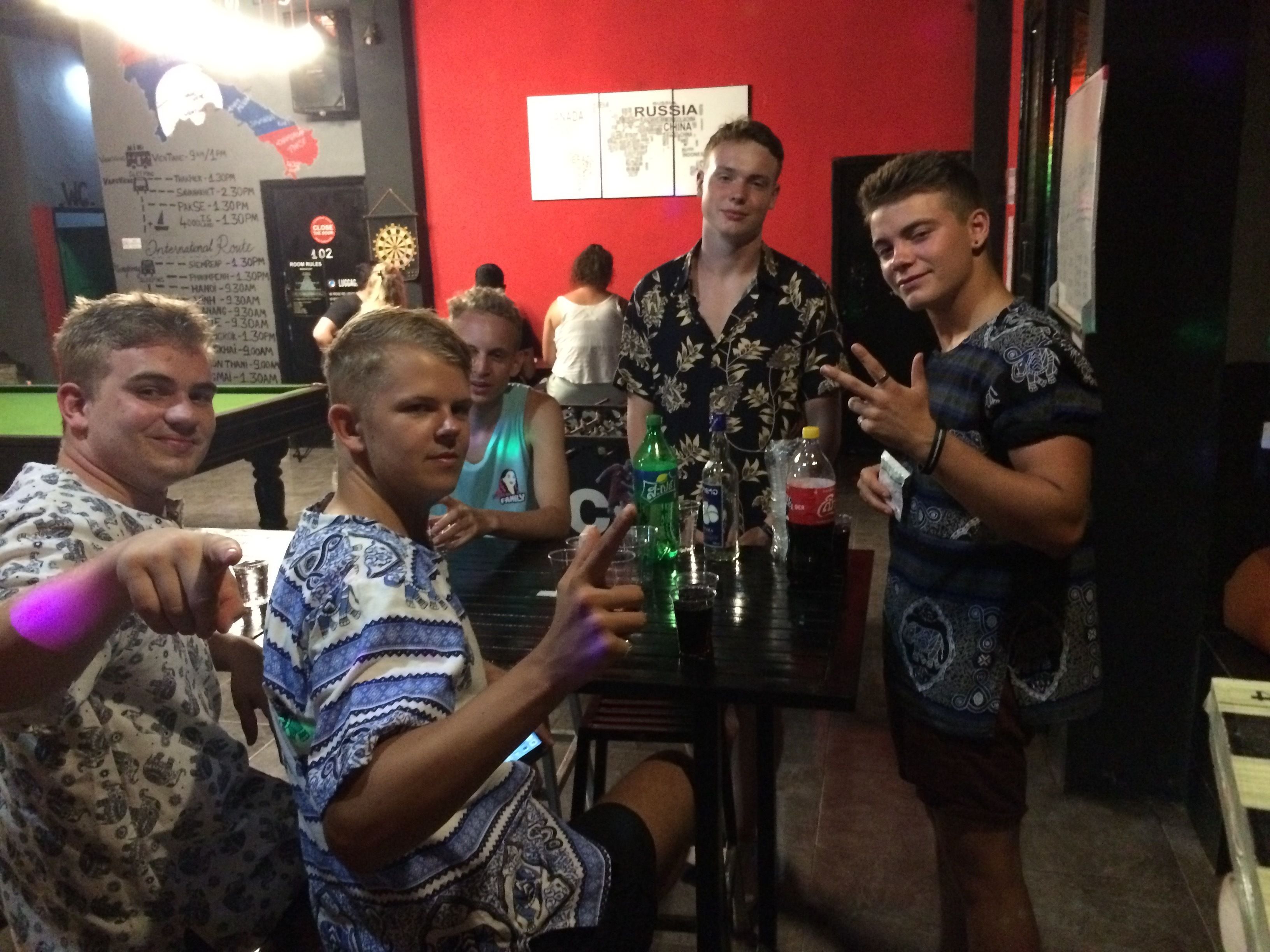 Un groupe de personnes se trouve dans une pièce faiblement éclairée, avec un mur rouge, un jeu de fléchettes et un tableau affichant le menu.