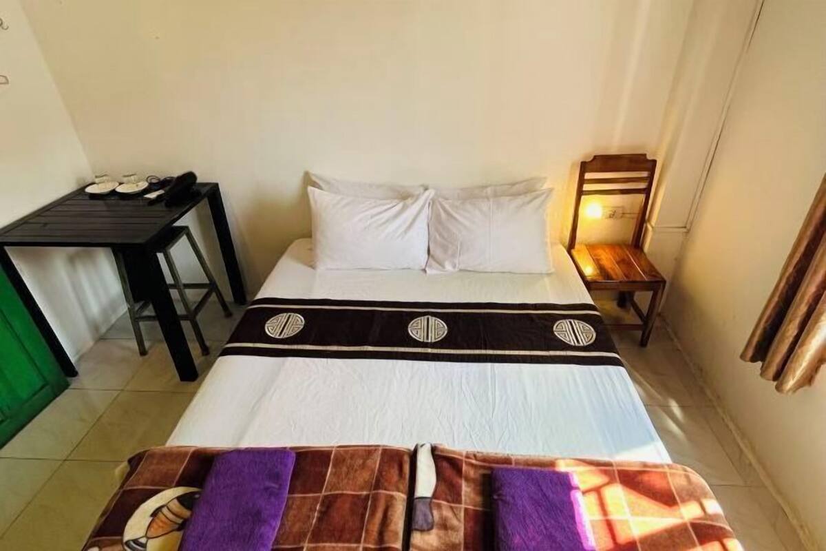 A hotel room with a bed, a small table with a chair, a wooden bench, and a wall-mounted heater.
