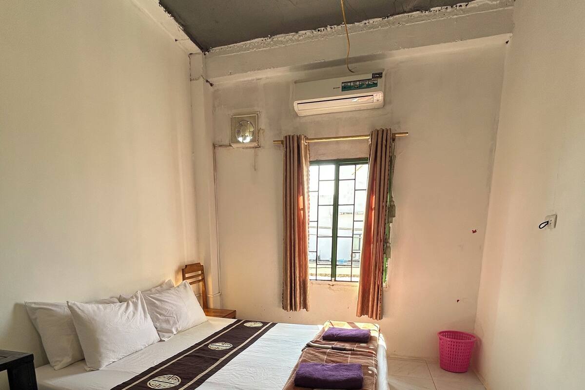 A small, clean room with a bed, a window with curtains, a ceiling fan, and a small air conditioning unit.