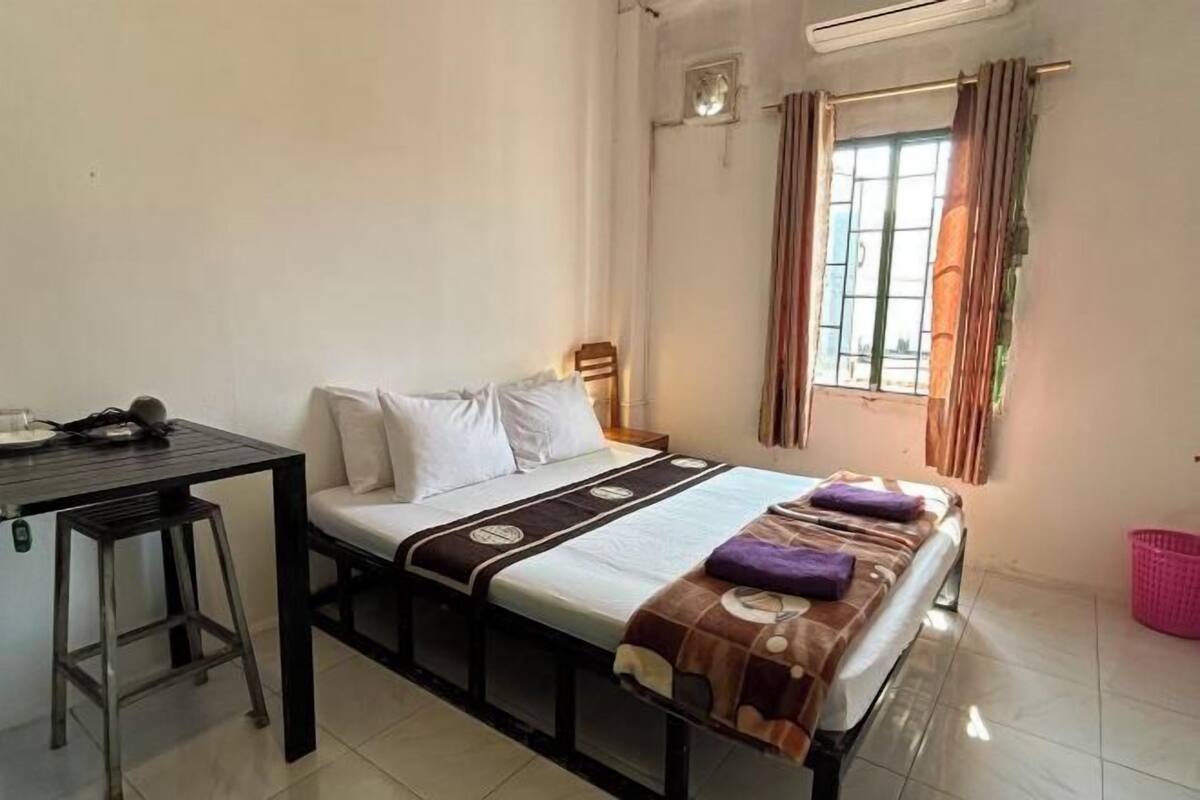 A single bed with a headboard, a small table with a chair, a window with curtains, and a wall-mounted air conditioning unit.