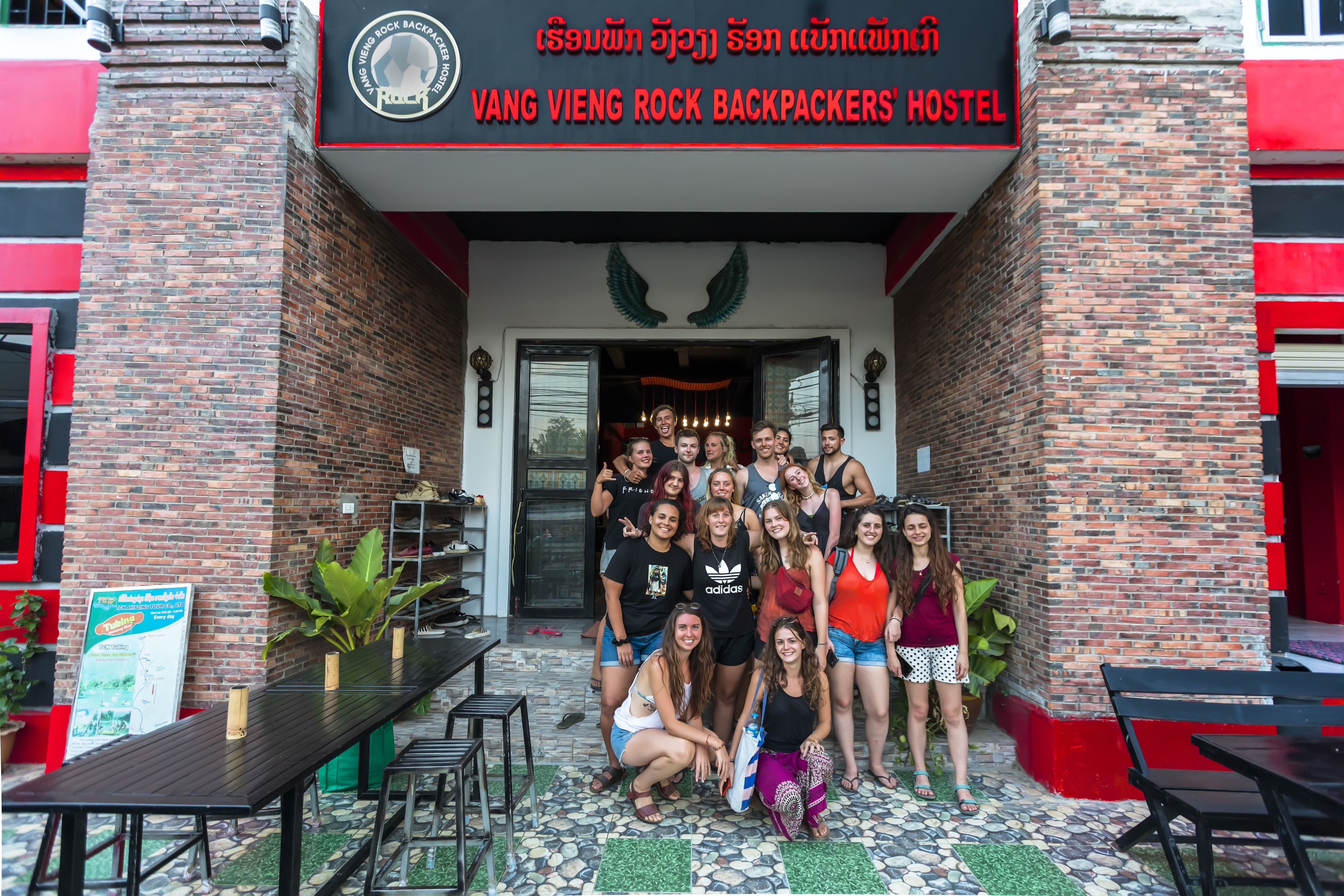 Un grup de persones fent-se una foto davant d'un hostal amb un cartell que diu "VANG VIEN ROCK BACKPACKERS HOSTEL".