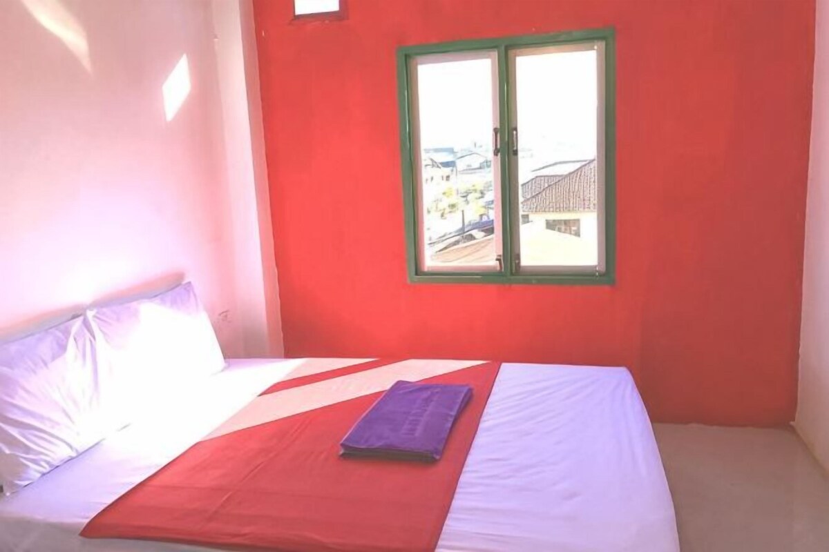 A bed with a white and red bedspread, a purple item on the bed, and a window with a green frame.