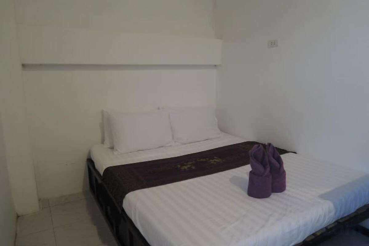 A single bed with a white mattress and a dark headboard, a white wall, and a small light switch.