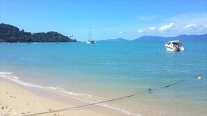 Una playa con agua cristalina, embarcaciones y un paisaje montañoso al fondo.