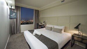 Habitación de hotel con una cama grande, televisión montada en la pared, escritorio con lámpara y vista nocturna de la ciudad.
