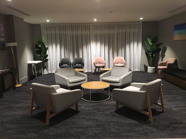 A modern lounge area with grey and pink upholstered chairs, a round coffee table, and a large window with sheer curtains. - Woodroffe Hotel (Southport)