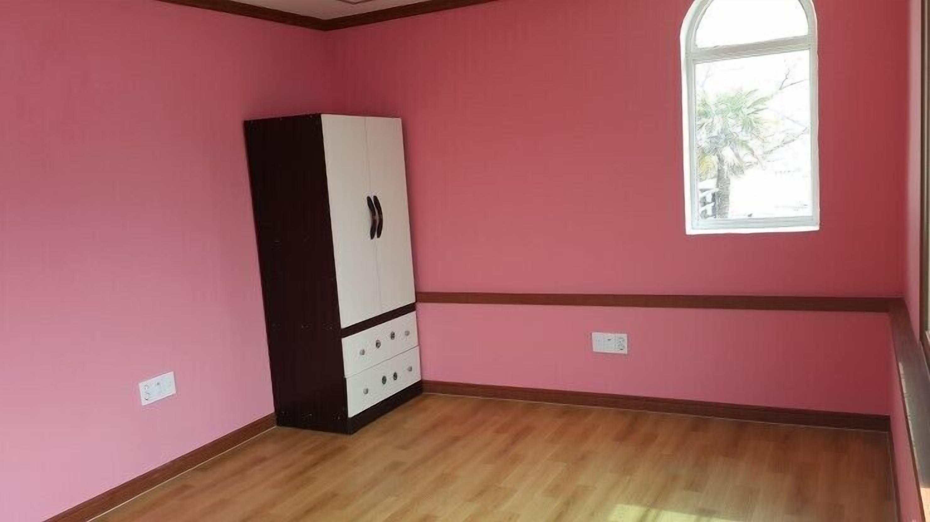 Una habitación con paredes rosas, suelo de madera, un armario blanco y negro, y una ventana con vistas a árboles.