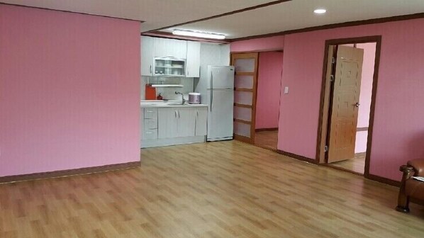 A room with pink walls, wooden flooring, a kitchen area with white cabinets and a refrigerator, and a wooden door. - Sirak Pension (Changwon)