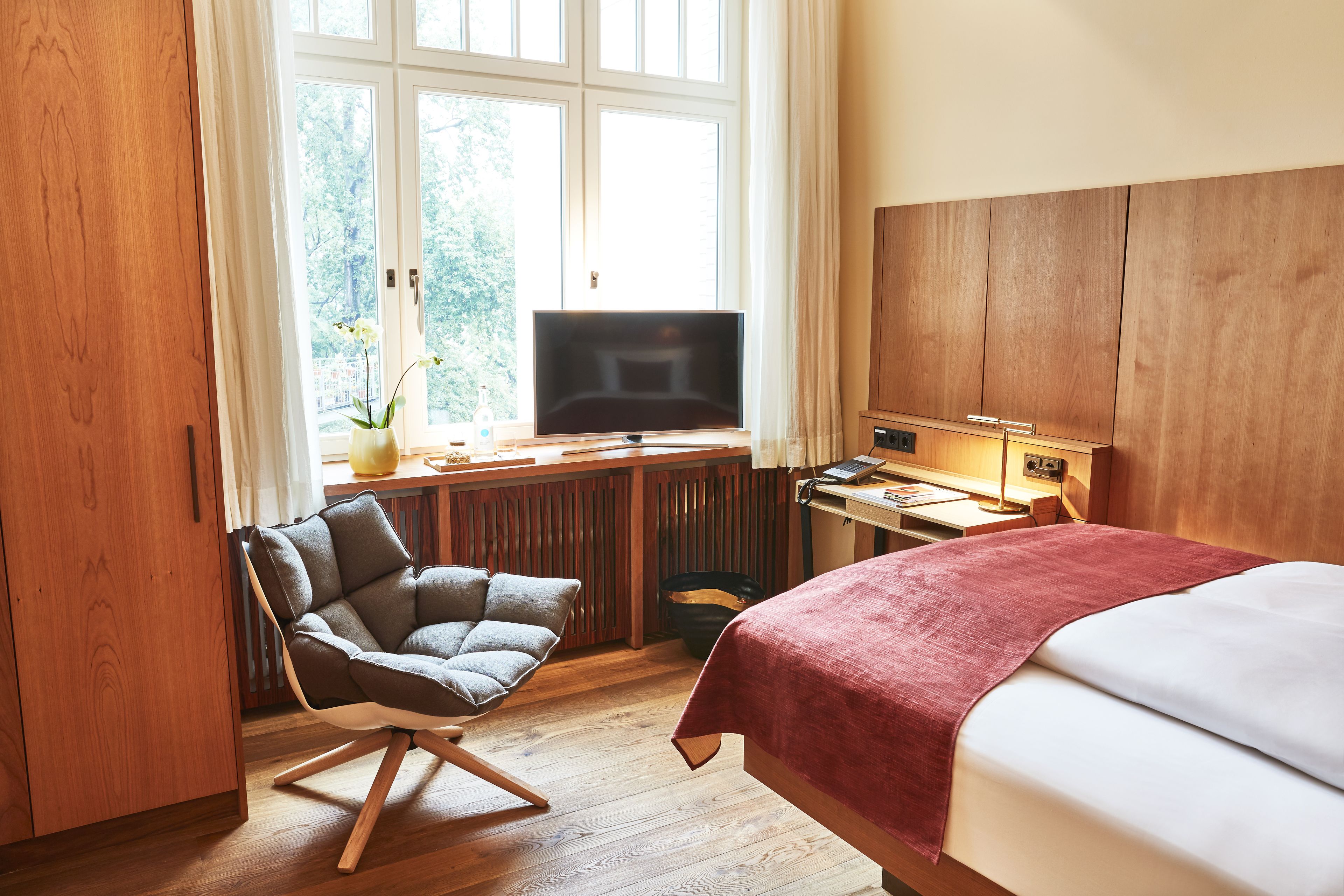 A hotel room with a bed, a desk with a television, a chair, and a window with a view of trees.