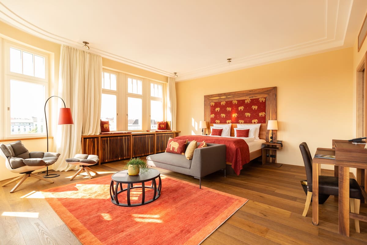 Ein Hotelzimmer mit Bett, Sofa, Esstisch und Stuhl. Ein Fenster mit Vorhängen, eine Lampe und eine Pflanze auf einem Tisch sind ebenfalls vorhanden.