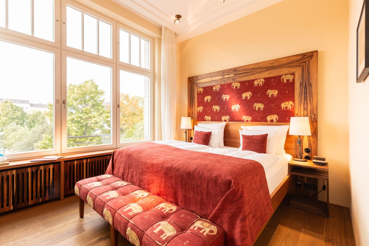 Ein Hotelzimmer mit einem großen Bett, zwei Nachttischen, einer Bank und einem Fenster mit Blick auf Bäume.