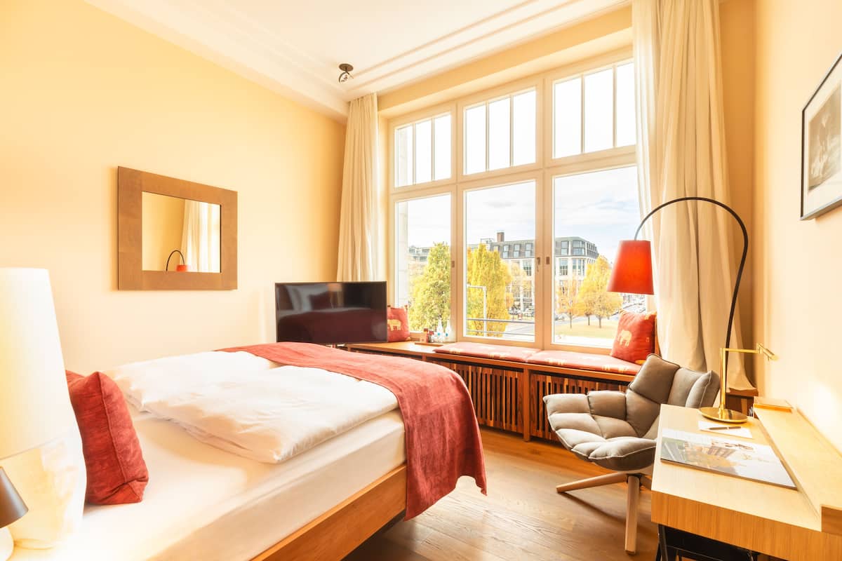 Ein Hotelzimmer mit Bett, Schreibtisch, Stuhl, Fernseher, Fenster mit Blick auf Gebäude und einer Lampe.