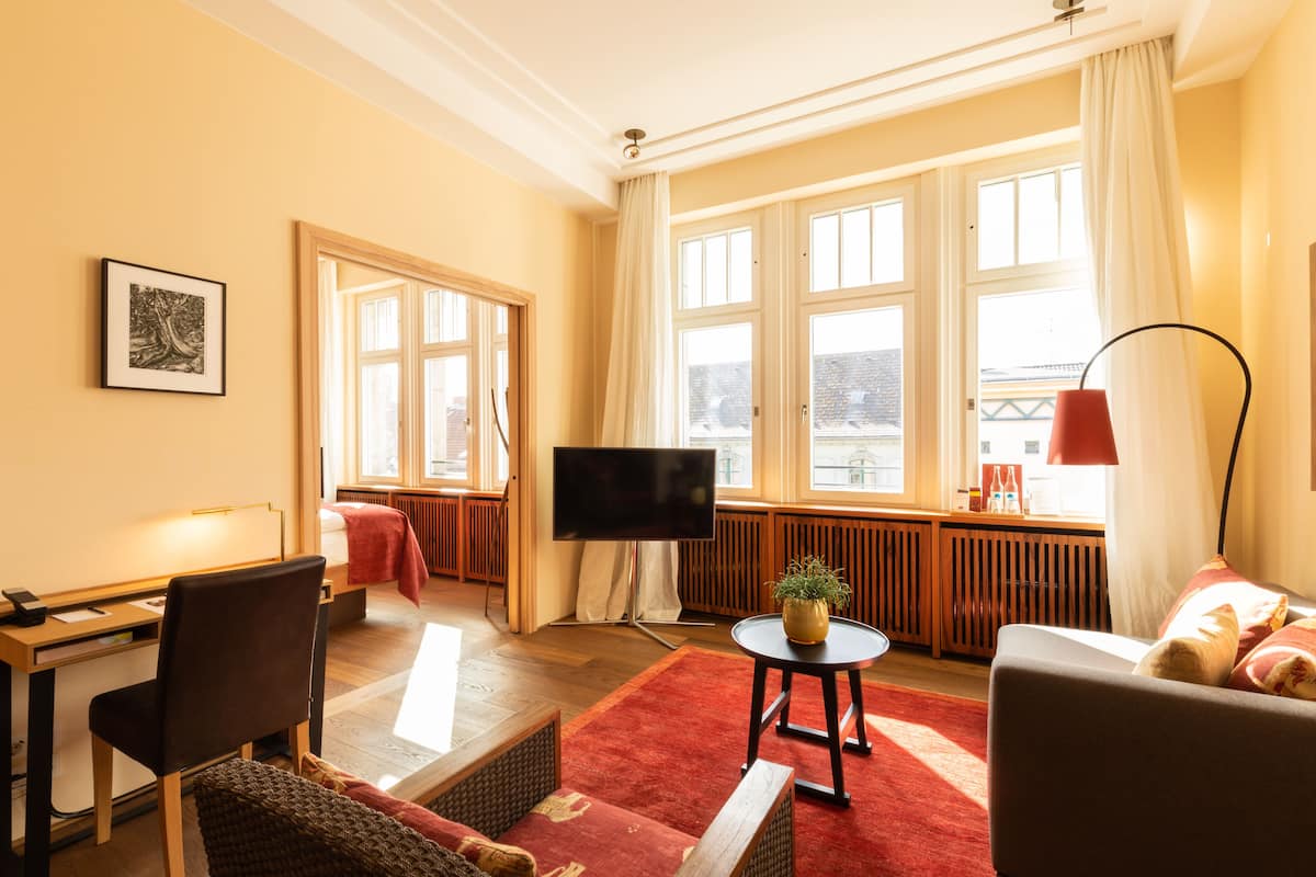 Ein helles Wohnzimmer mit Fernseher, einer Couch, einem Sessel und einem roten Teppich.