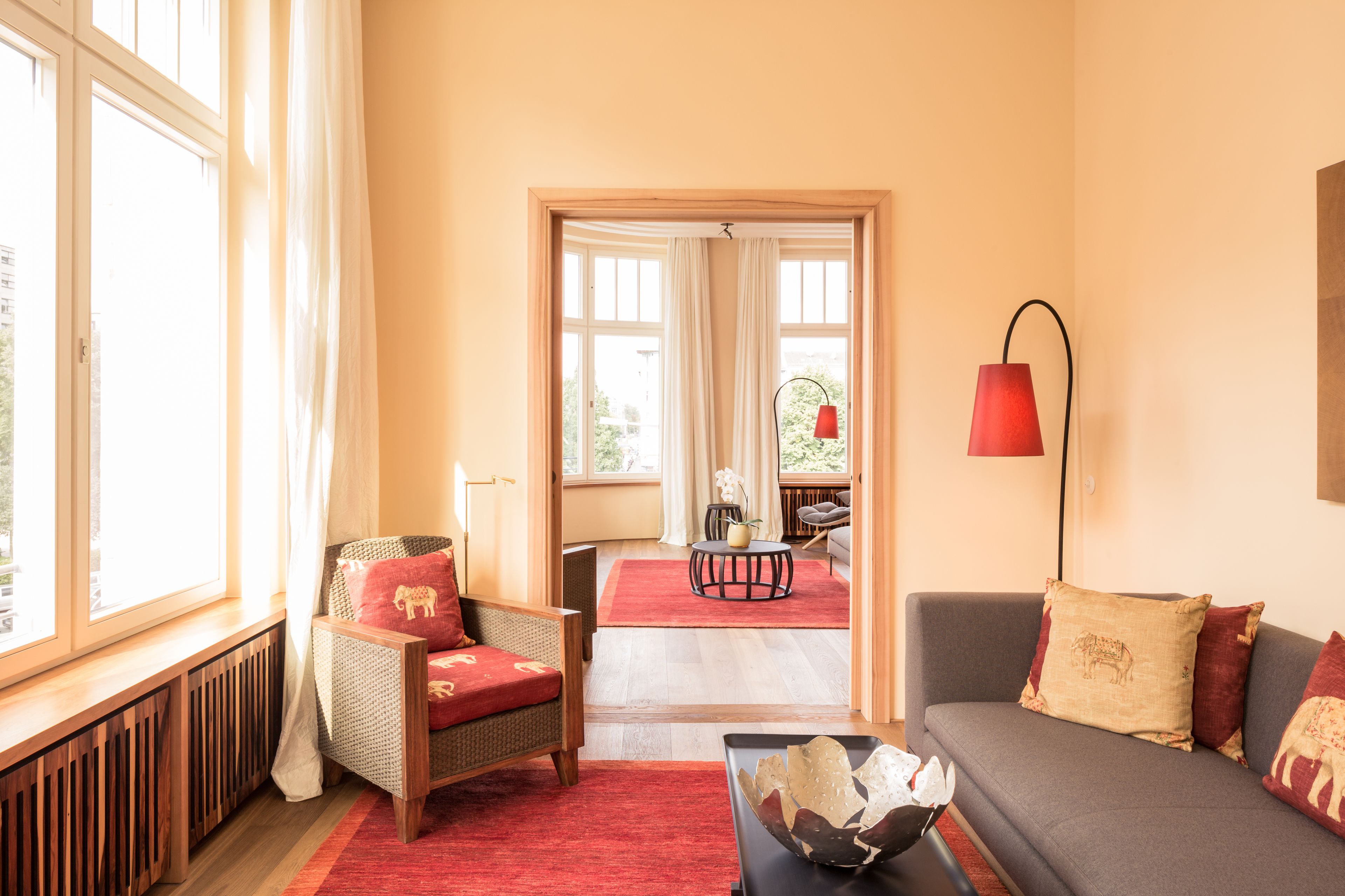 A living room with a sofa, armchair, and a red rug. There is a window with curtains, a lamp, and a view of an outdoor area.