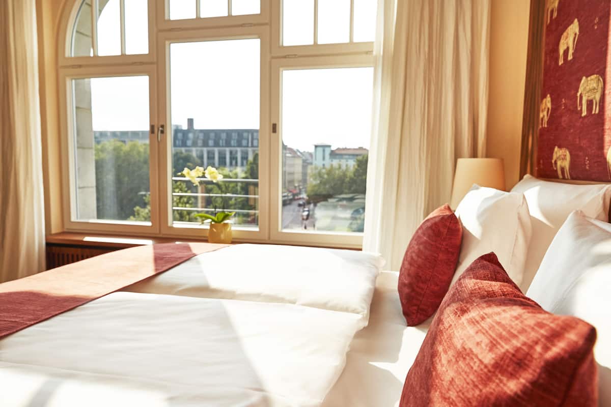Ein Hotelzimmer mit einem großen Bett, einem Fenster mit Blick auf Gebäude und Grünflächen, einem Blumentopf und einer Wand mit einem gemusterten Design.