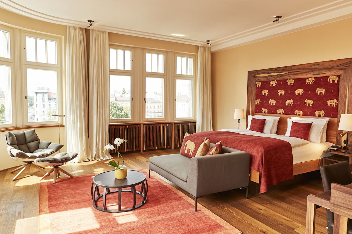 Ein Hotelzimmer mit einem großen Bett, einem Sofa, einem Sessel, einem kleinen Tisch und Blick auf die Stadt durch große Fenster.