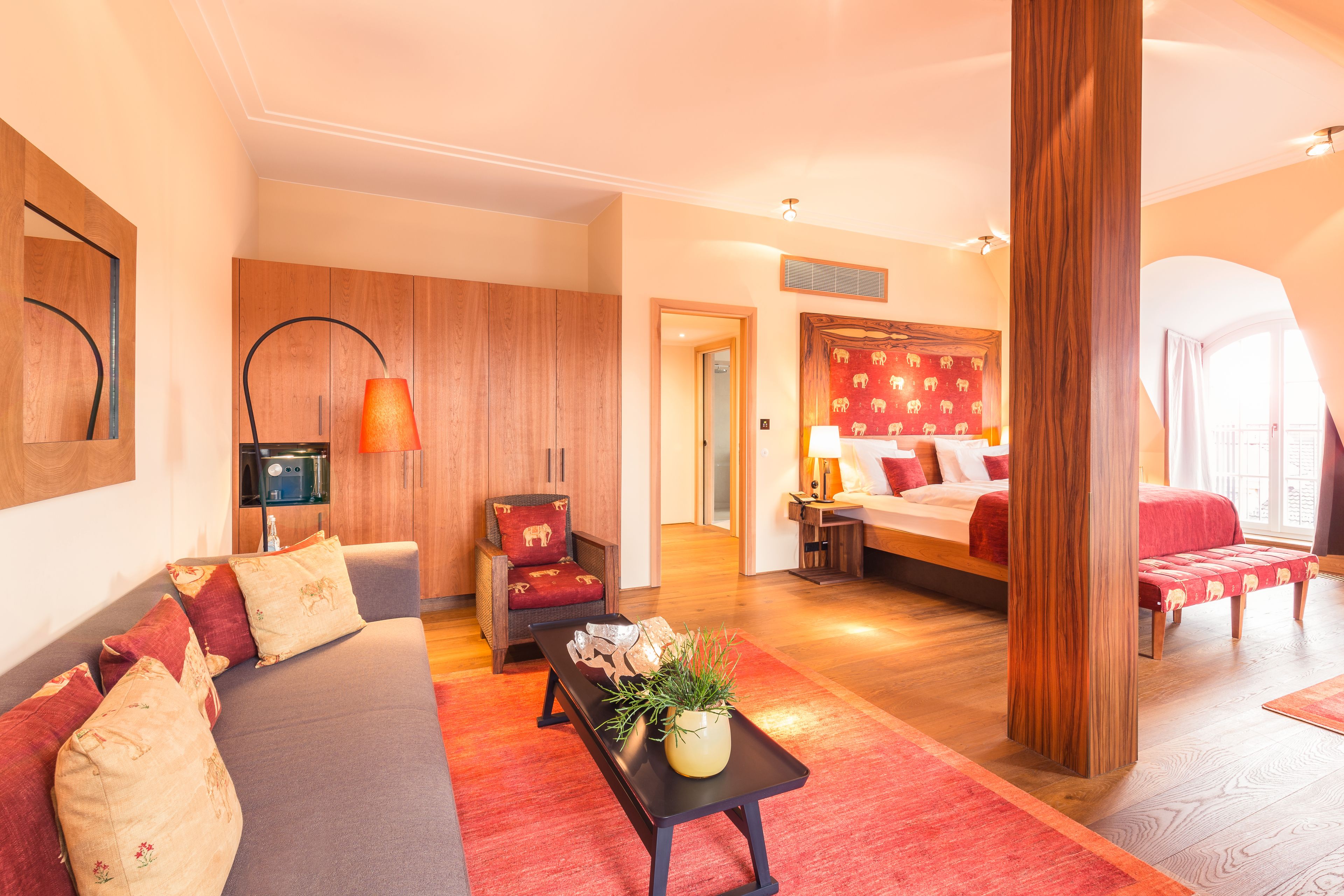 Een moderne hotelkamer met een bank, een houten accentmuur, een bed met rode hoofdbord en een groot raam.