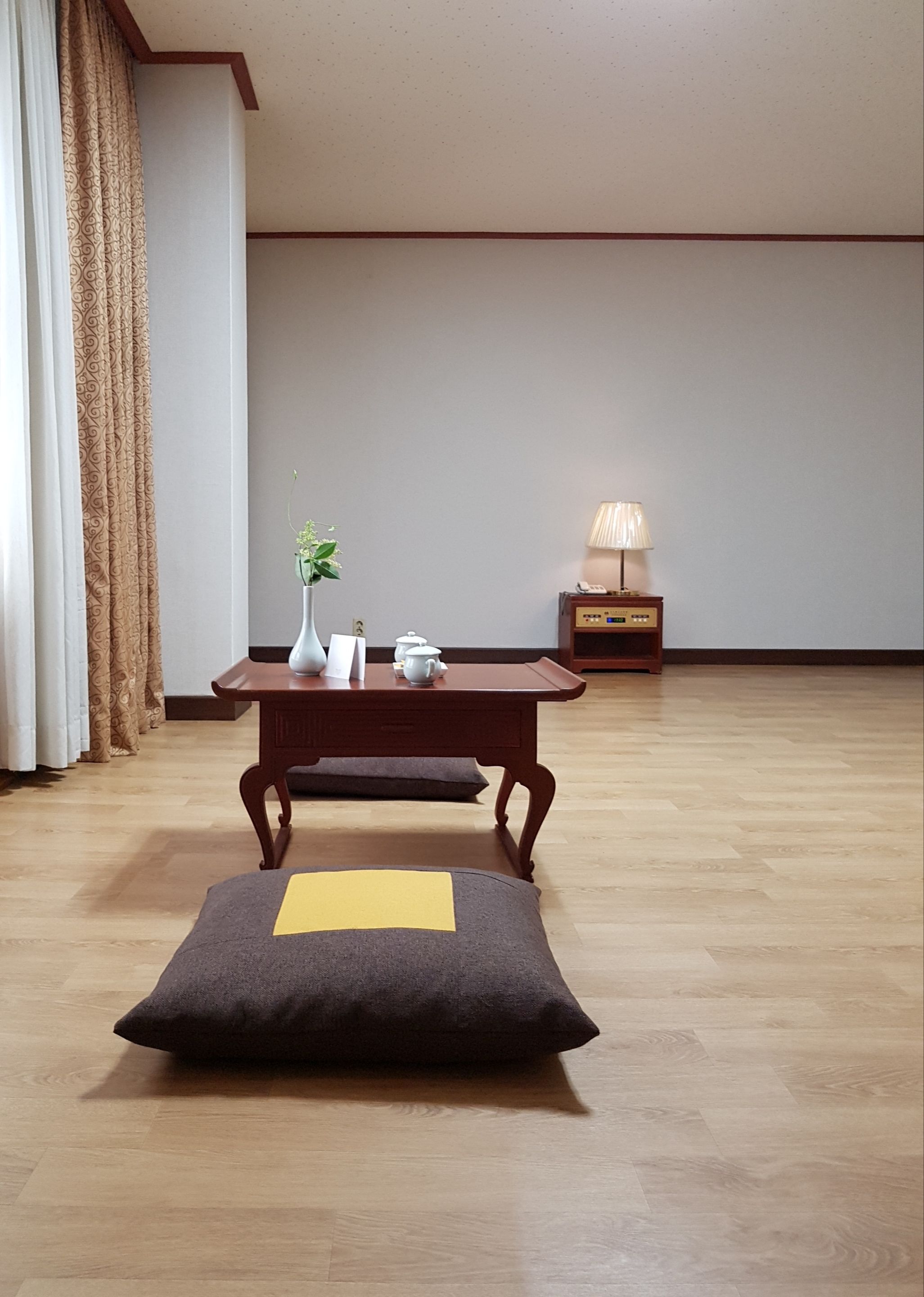 Una habitación con piso de madera, una mesa pequeña con un florero y una tetera, una lámpara y un cojín con un detalle amarillo.