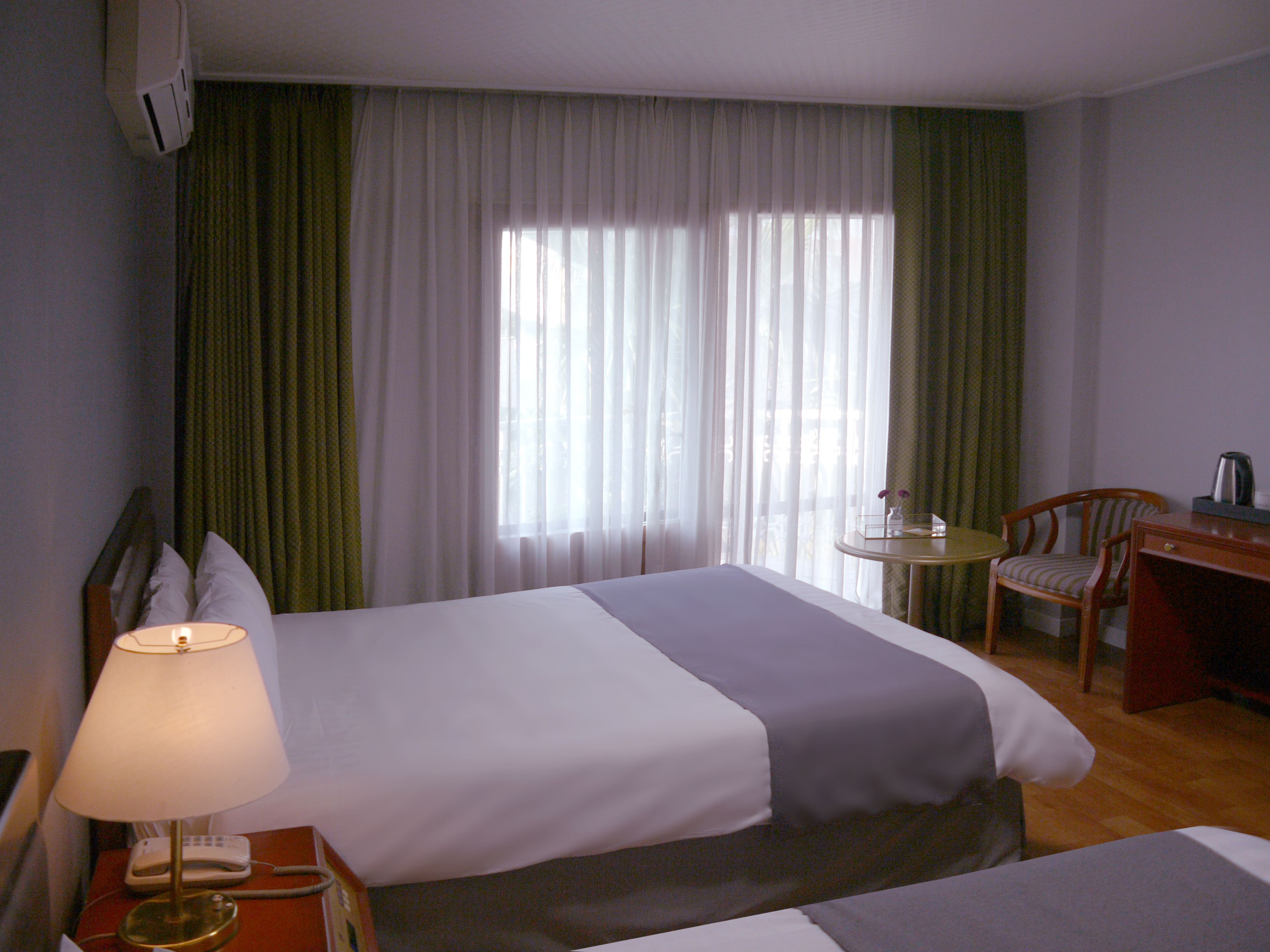 Een hotelkamer met twee bedden, een bureau, een stoel, een lamp en een raam met dunne gordijnen.