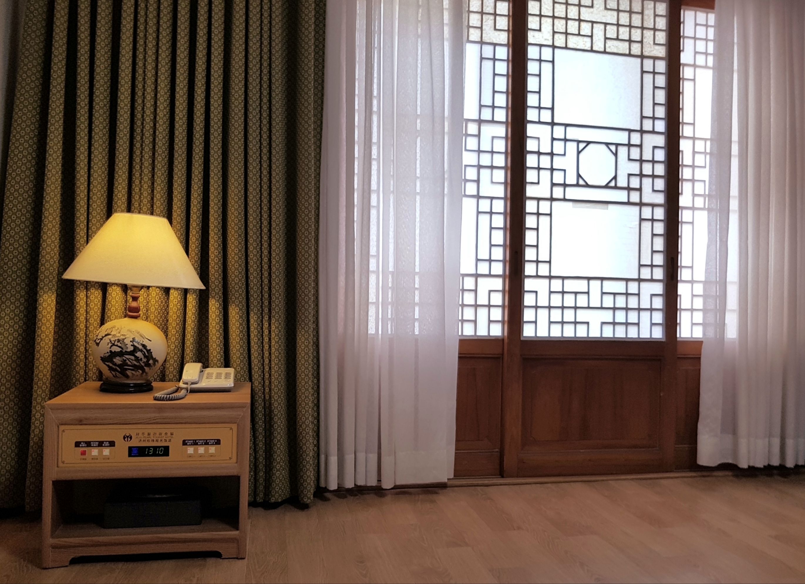 A room with a wooden nightstand, a lamp, a telephone, and a digital clock.
