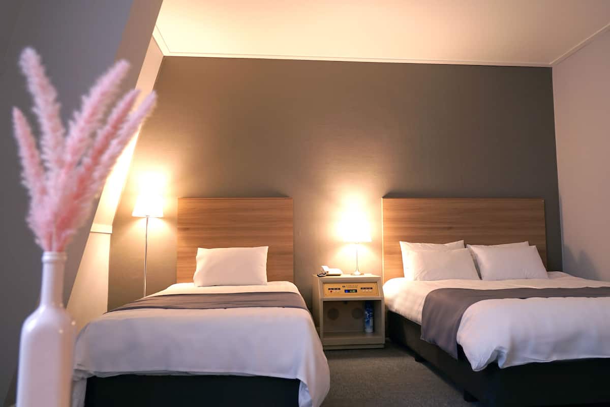 Una habitación de hotel con dos camas, una mesita de noche con lámpara y un jarrón con flores rosadas.