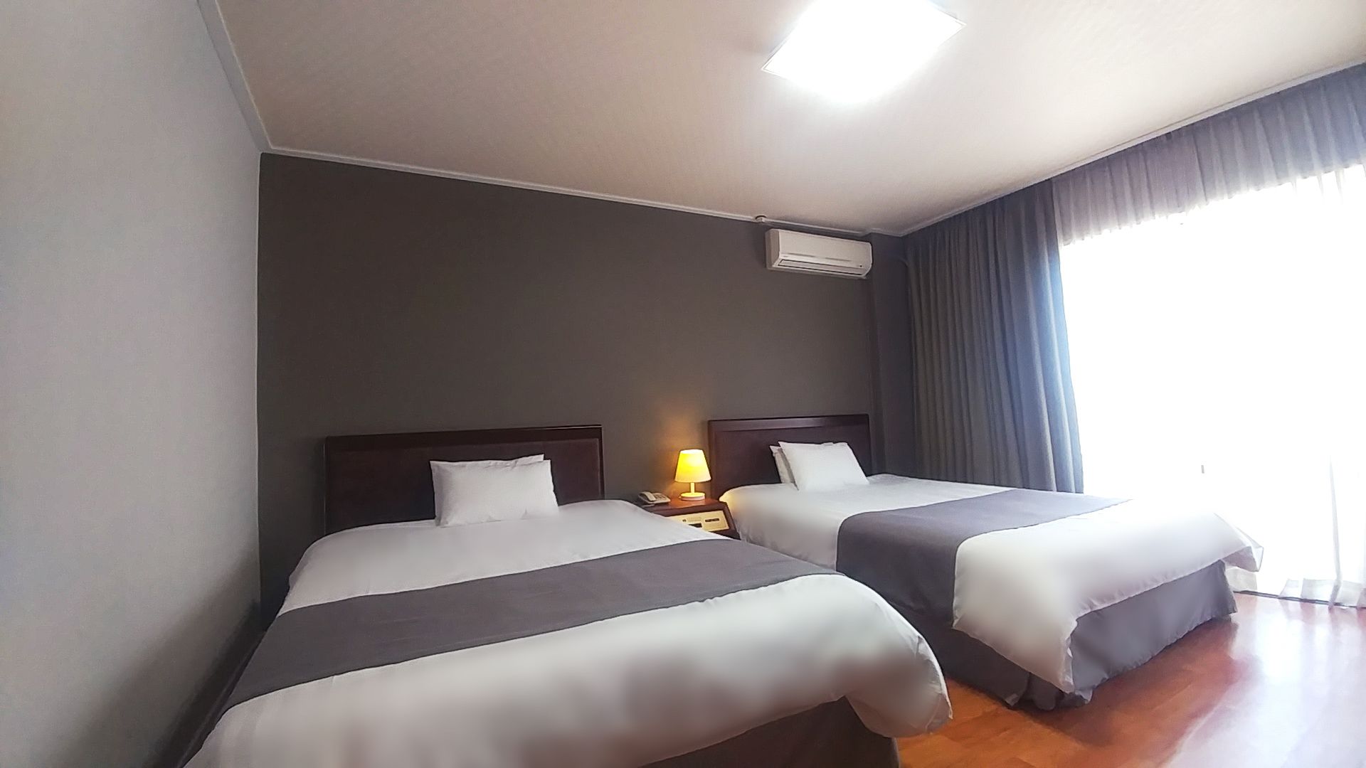 A hotel room with two beds, a nightstand with a lamp, an air conditioning unit, and a large window with curtains.