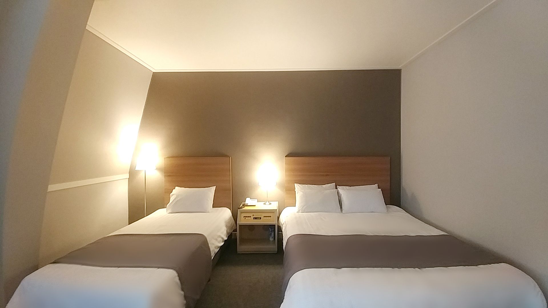 A hotel room with two single beds, each with a wooden headboard, a bedside table, and a lamp.