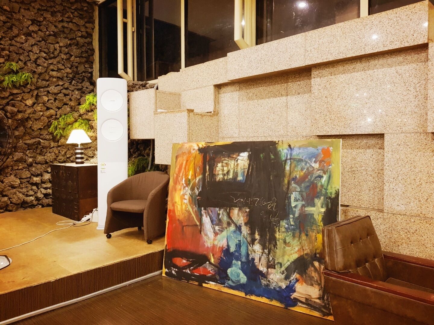 Quarto com parede de pedra, uma cadeira moderna, uma mesa com luminária e um quadro abstrato colorido.