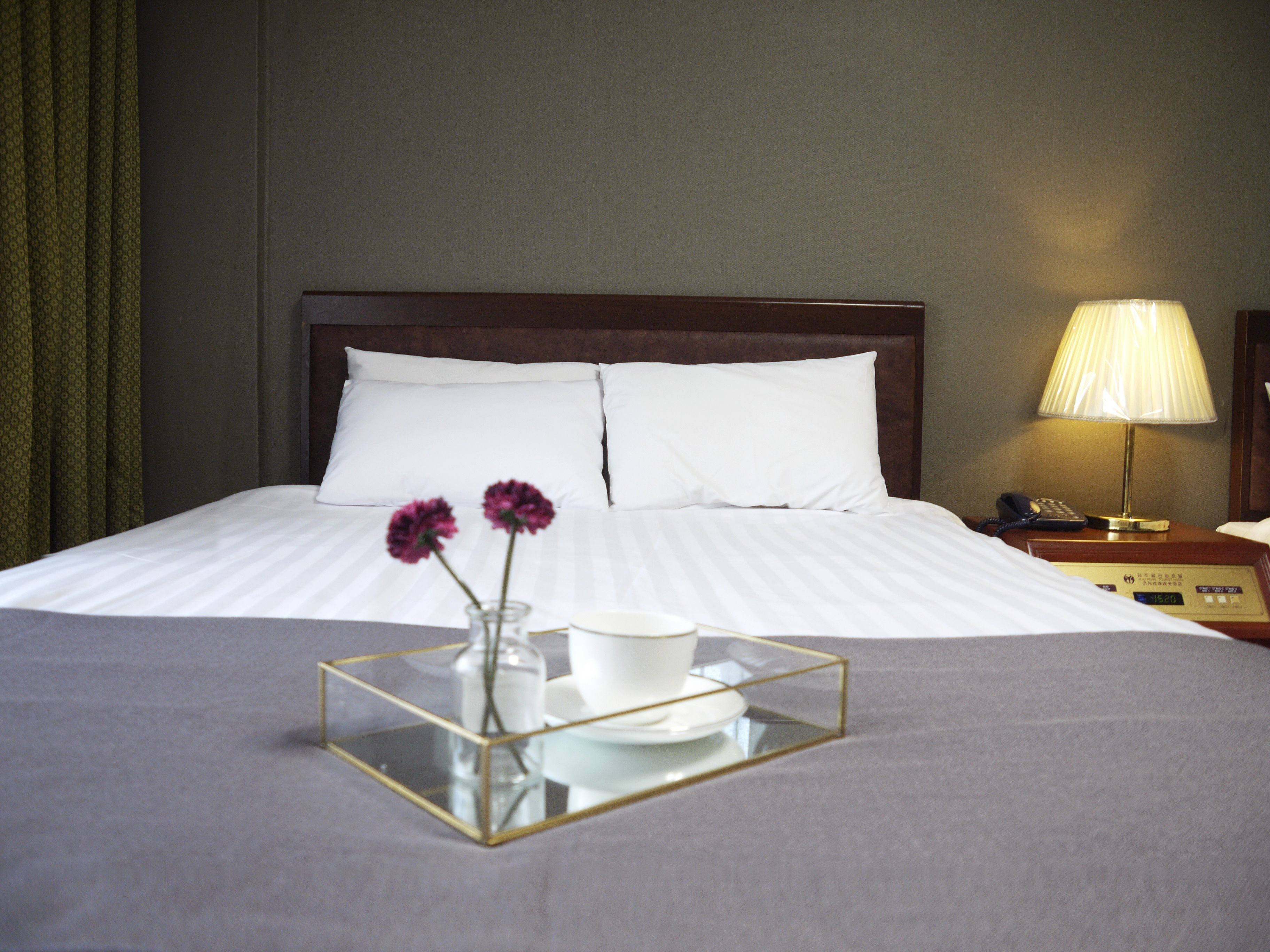 Una cama bien tendida con sábanas blancas, una mesita de noche con lámpara y una bandeja con una taza y flores.