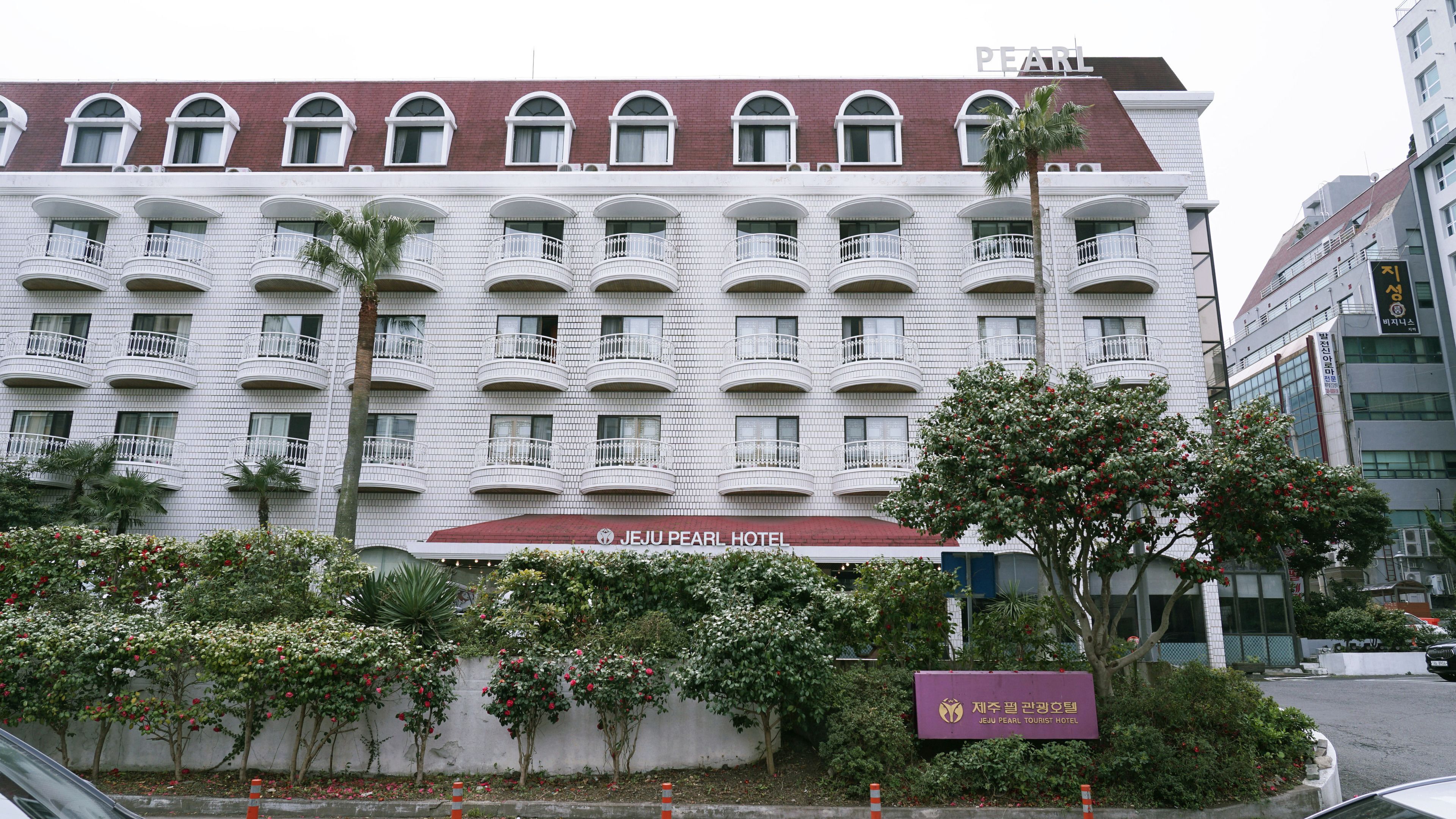 A multi-story hotel with a red roof and white facade, featuring balconies and a sign for Jeju Pearl Tourist Hotel.