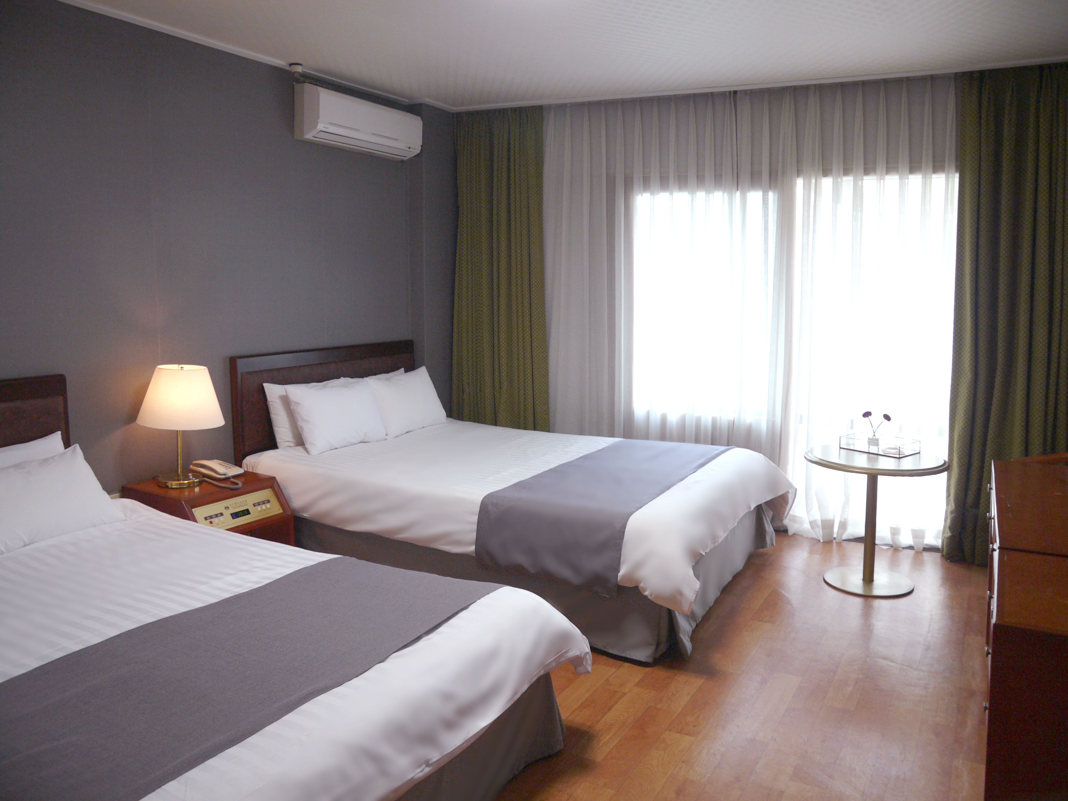 A hotel room with two beds, a bedside table with a lamp, a small table with a vase, and a window with curtains.