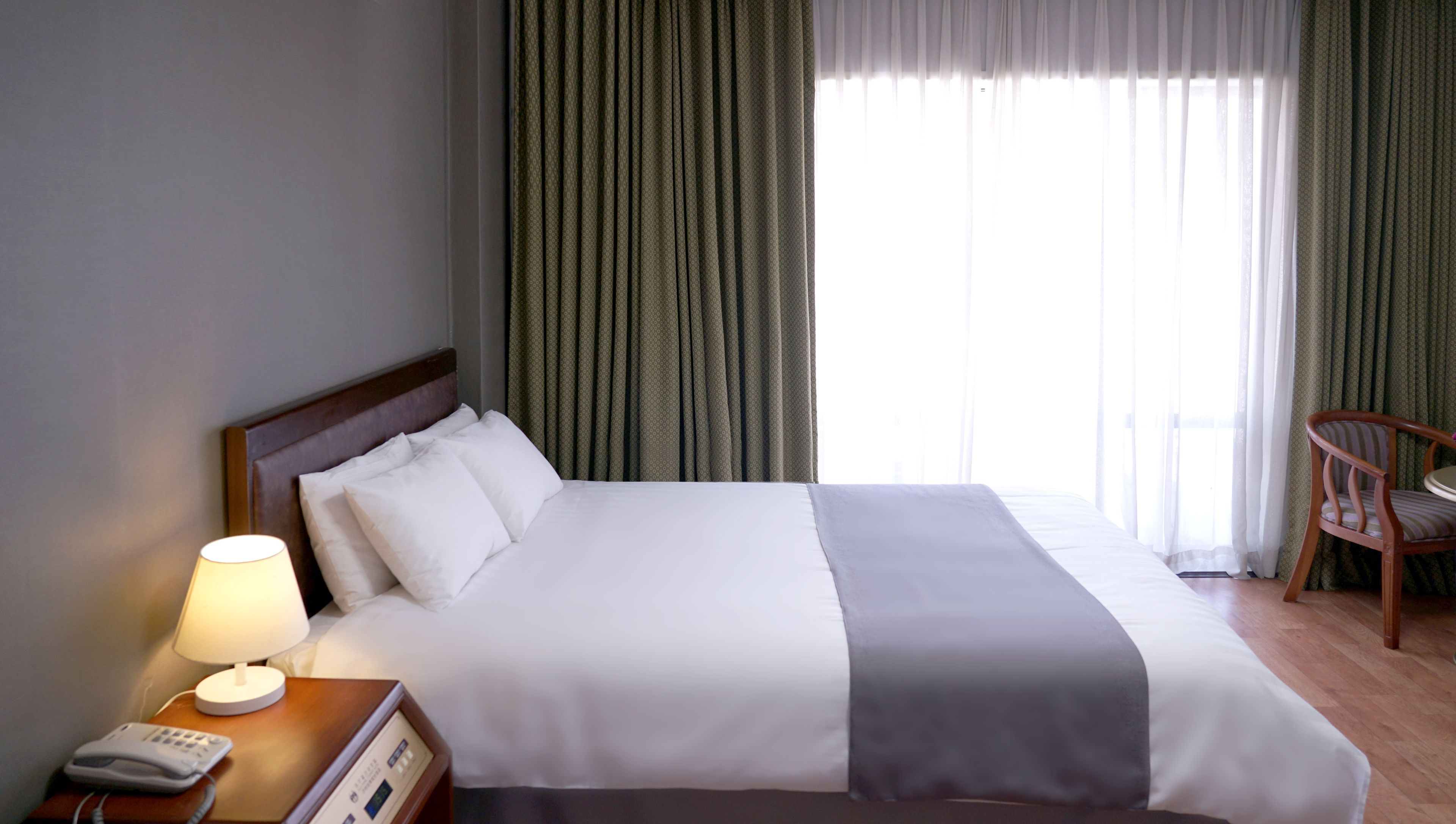 Een hotelkamer met een bed, bedlampje, telefoon en stoel bij een raam met gordijnen.