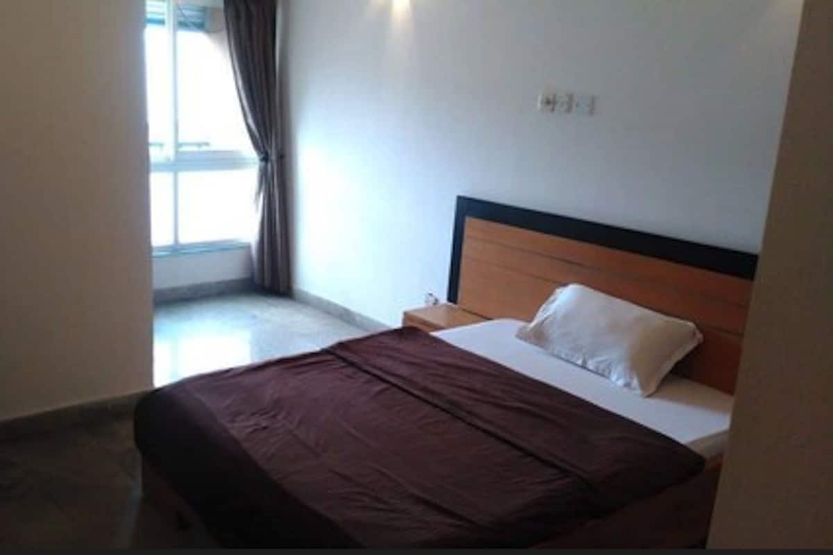 A hotel room with a bed, a nightstand, a window with curtains, and a wall-mounted light switch.