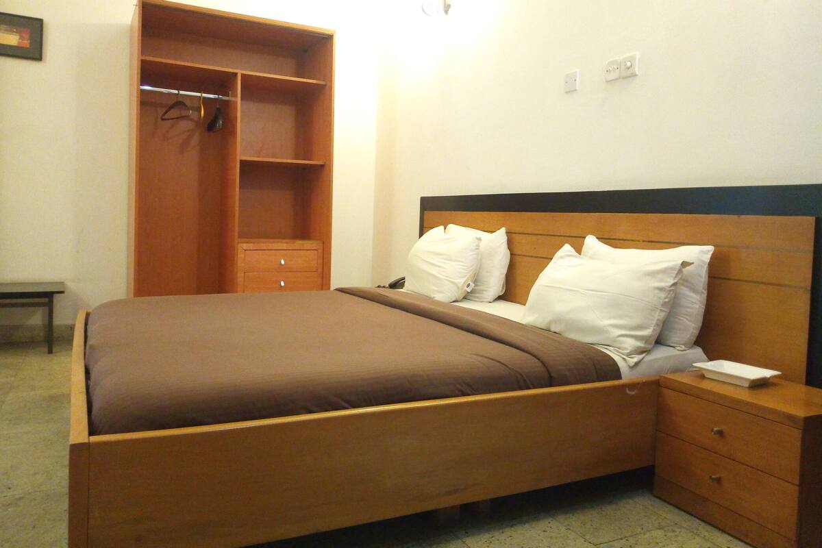 A bedroom with a bed, a nightstand, a wooden wardrobe, and a wall-mounted light.