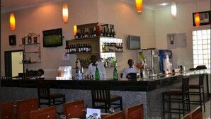 A bar area with a counter, bar stools, and a television screen. There are shelves with bottles and a refrigerator. - Karma Hotel (Lagos)
