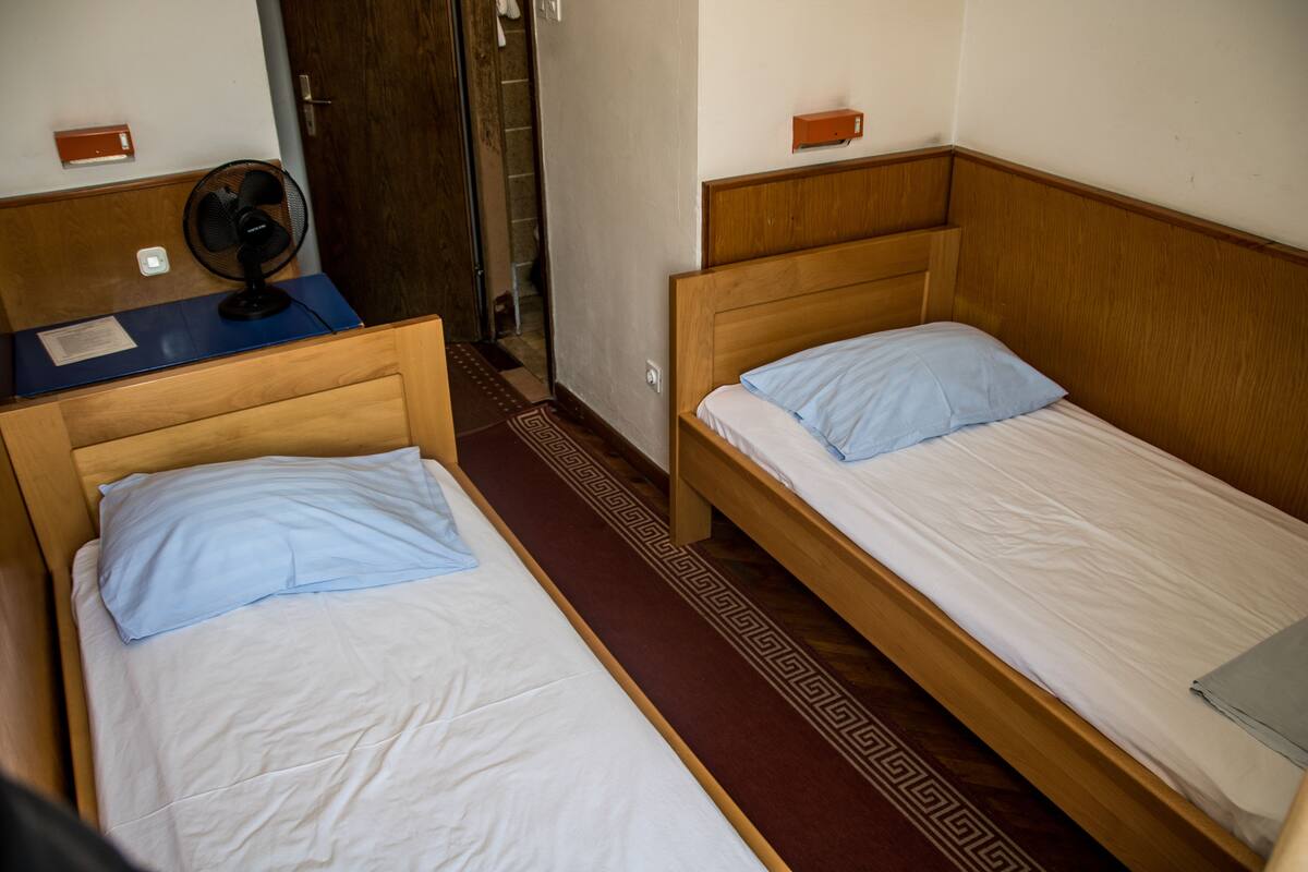 Zwei Einzelbetten mit Holz Kopfteilen, ein kleiner Schreibtisch mit Ventilator und ein gemustertes Teppich.
