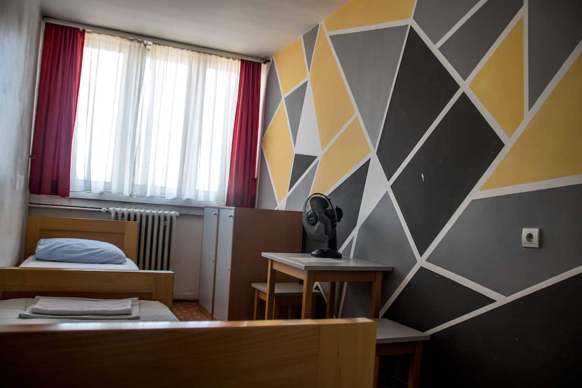 Ein Hotelzimmer mit einem Bett, einem Schreibtisch, einem Sessel, einem Fenster mit Vorhängen und einer Wand mit geometrischem Muster.
