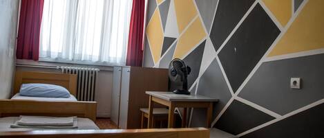 A hotel room with a bed, a desk, a chair, a window with curtains, and a geometric-patterned wall.