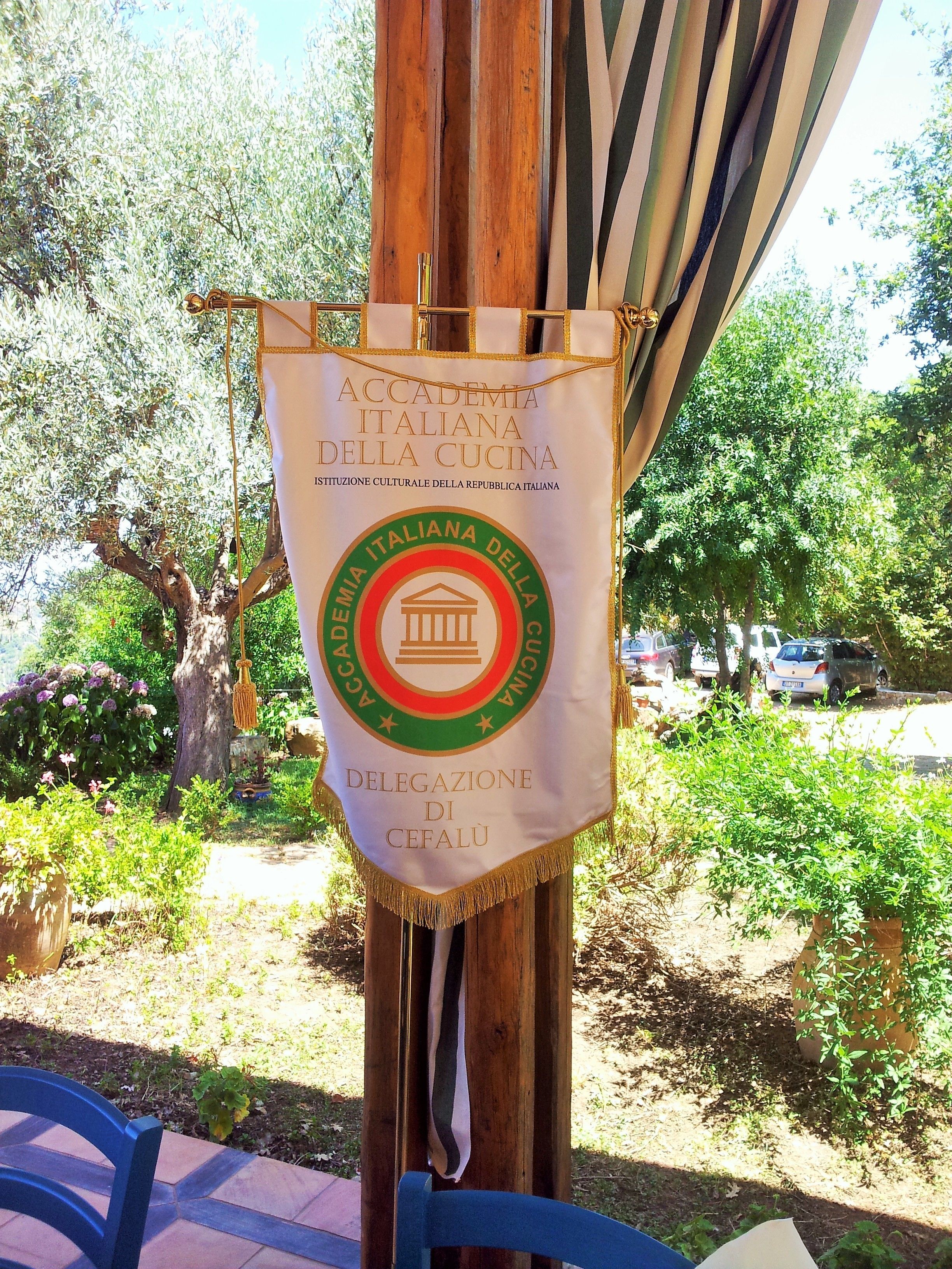 A banner hanging from a wooden post with the text "Accademia Italiana della Cucina" and "Delegazione di Cefalù".
