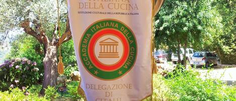 A banner hanging from a wooden post with the text "Accademia Italiana della Cucina" and "Delegazione di Cefalù".
