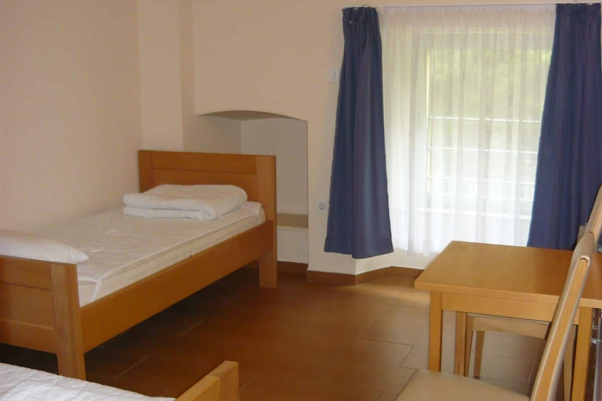 A room with two wooden beds, a wooden table with a chair, and a window with curtains.