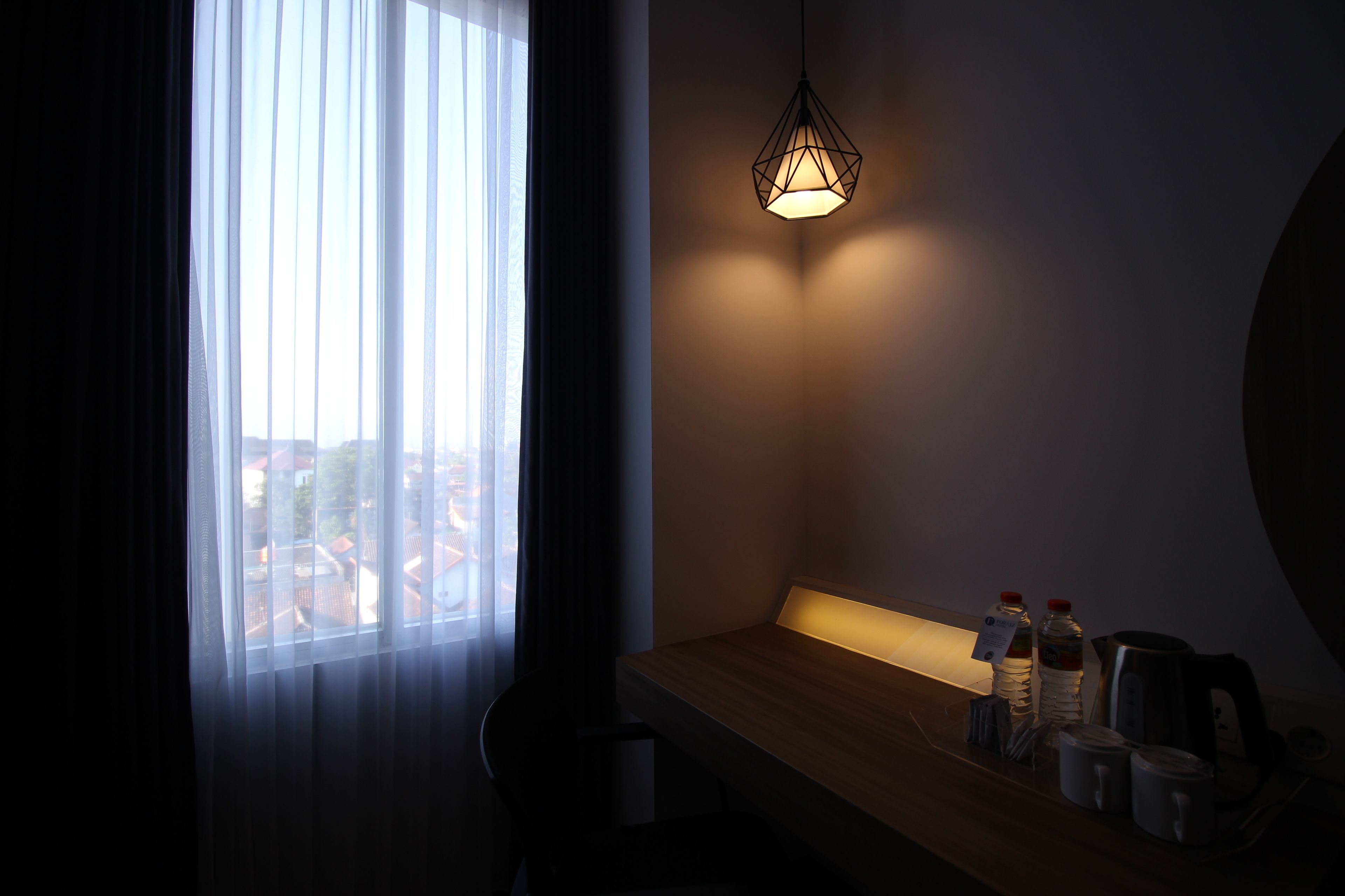 Una habitación tenuemente iluminada con una ventana, un banco de madera y una mesa con botellas y una tetera.